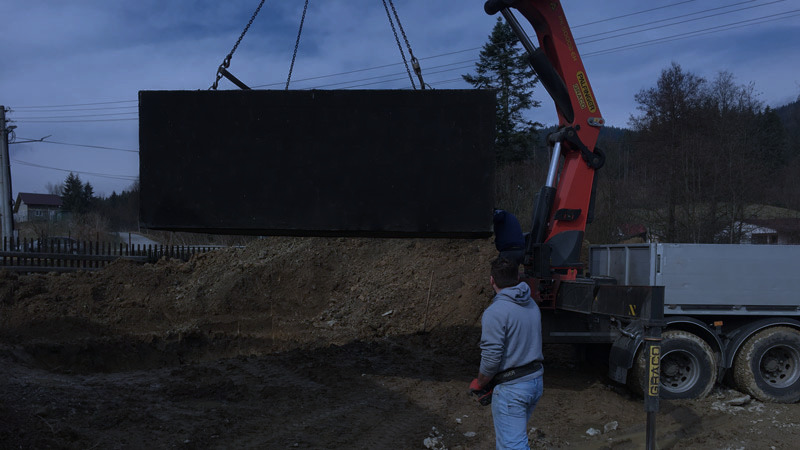 Fotografia zachytáva profesionálne osadenie betónovej žumpy do pripraveného výkopu pomocou vozidla s hydraulickou rukou. Obrázok prezentuje reálnu montáž betónovej nádrže priamo u zákazníka a zdôrazňuje dovoz aj osadenie v cene po celej SR.
